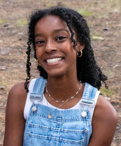 Hannah Barine wears a white tank and jean overalls and smiles at the camera