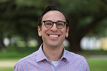 Daniel Barish wears a checked collared shirt and faces the camera smiling