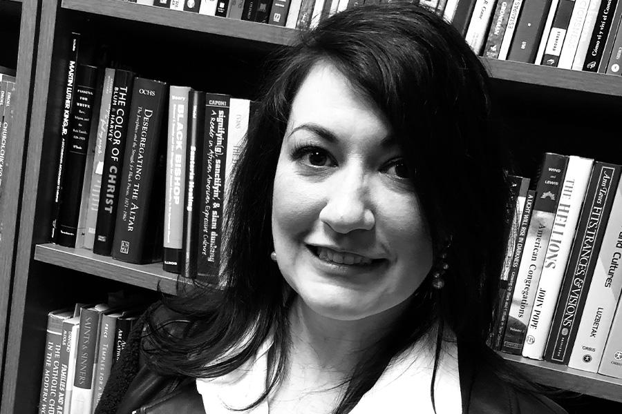 Dr. Maggie Elmore wears white collared shirt and dark blazer and smiles in front of bookshelf