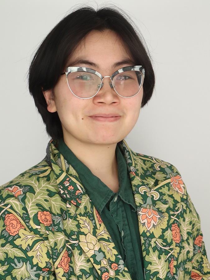Misa Nguyễn wears glasses and a dark green shirt and green floral blazer and smiles at the camera