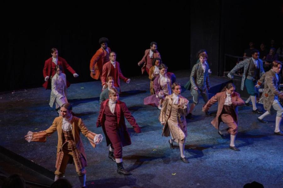 Male and female students dance onstage wearing 18th century men's suits