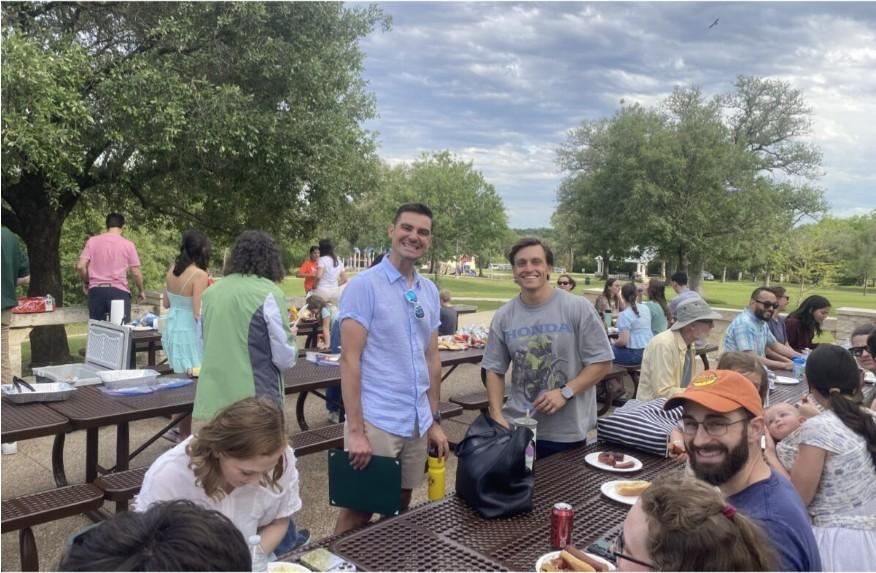 Professors and students in casual clothing stand in a park at picnic tables, smiling and eating