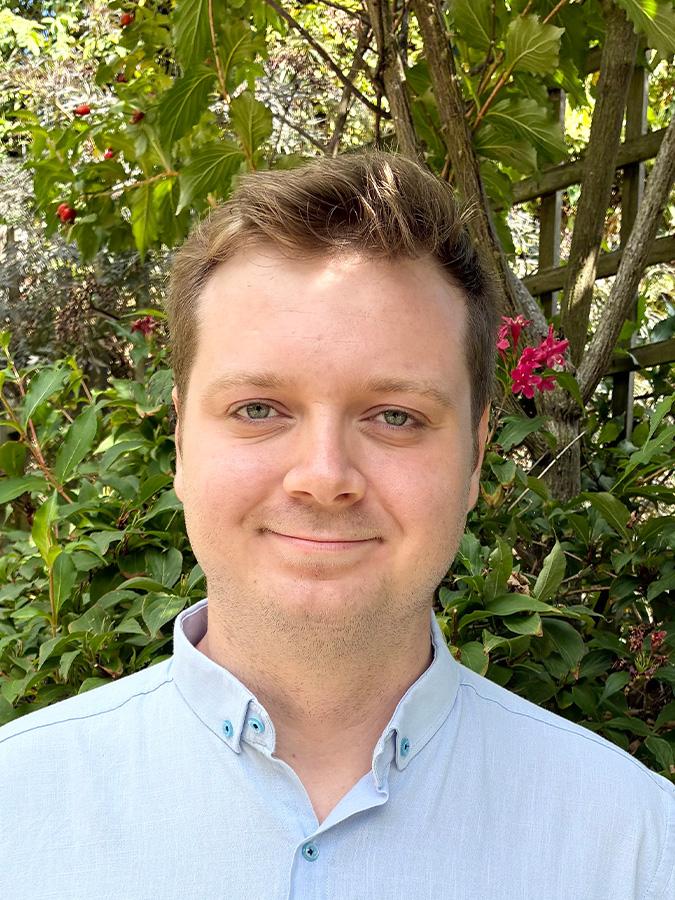 Headshot of Oliver Ind standing outside in blue shirt, smiling at the camera.