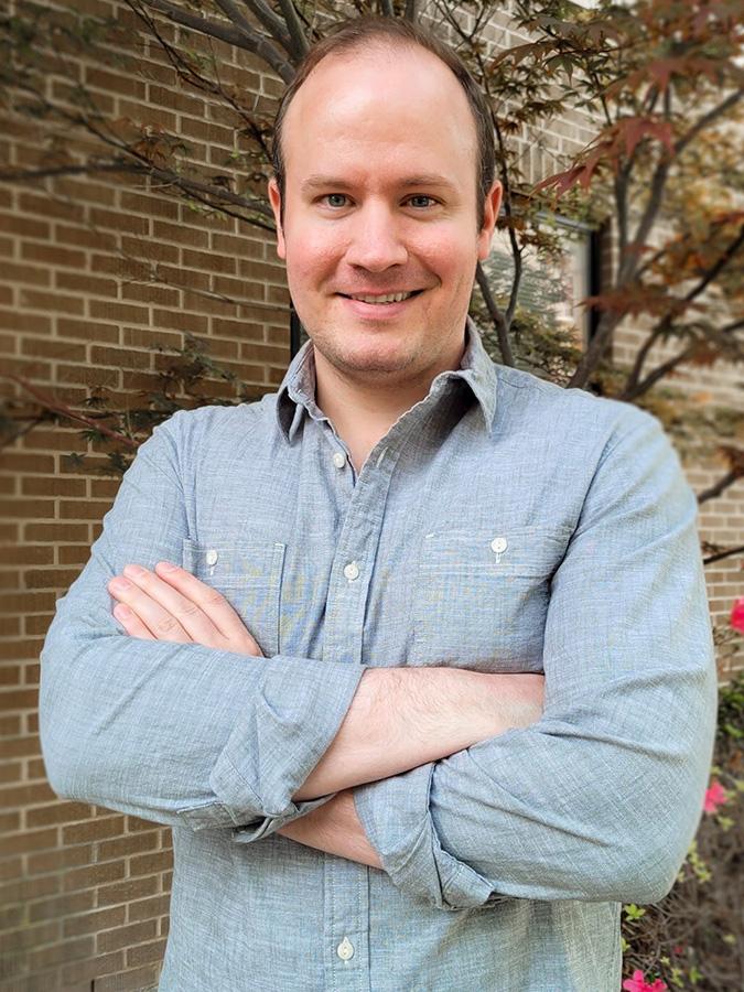 Joe Walton wears a blue long-sleeved shirt and crosses his arms, smiling at the camera.